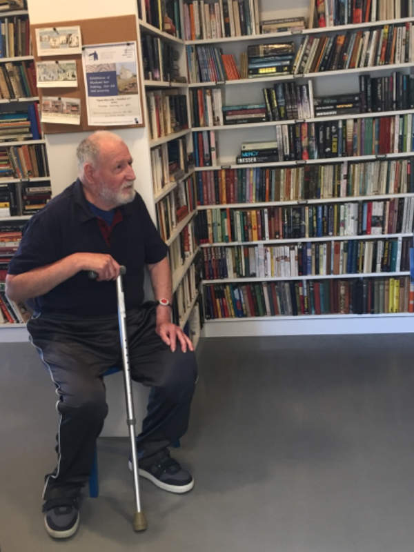 A man with a walking stick is sitting on a chair by well-stocked bookshelves