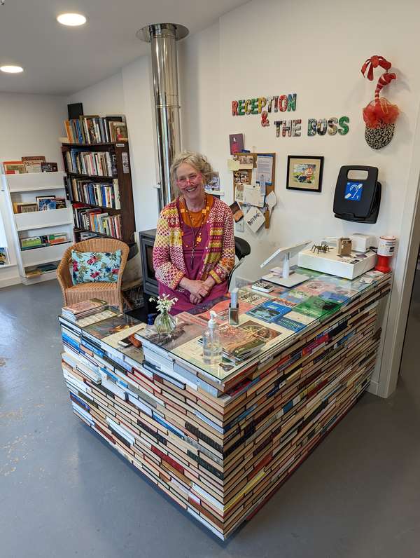 Sarah standing behind a desk made out of books, with the sign on the wall which reads 'Reception and the boss'